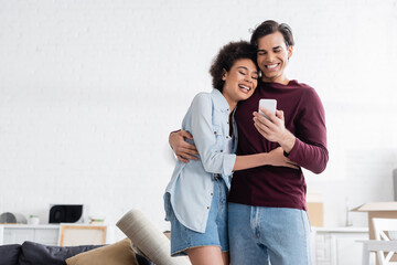 happy man using smartphone and hugging smiling african american girlfriend