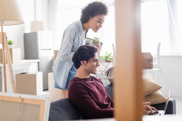 cheerful african american woman holding basket near smiling boyfriend using laptop