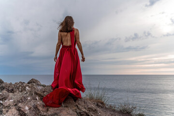 Fototapeta na wymiar A woman in a red dress stands above a stormy sky, her dress fluttering, the fabric flying in the wind.