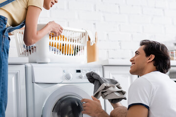 smiling man loading washing machine near cheerful african american girlfriend with basket of dirty laundry