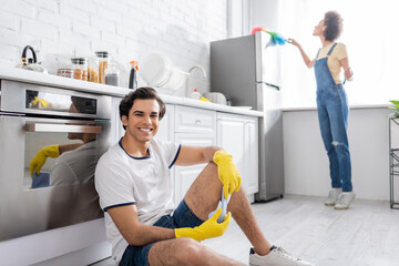 happy young man sitting near kitchen cabinet near curly african american woman cleaning fridge with dust brush in kitchen