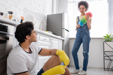 curly african american woman holding dust brush and looking at blurred boyfriend in rubber gloves