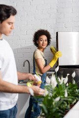 smiling african american woman holding sponge near fridge near blurred man watering plants in kitchen
