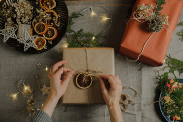 Female hands holds Christmas present wrapped in kraft paper. DIY Christmas eco friendly packaging for holiday Christmas gifts. Table top view.