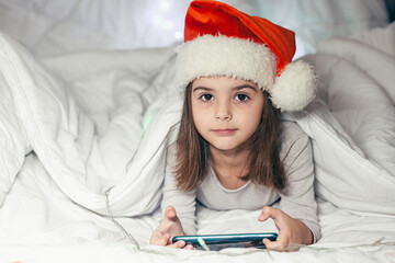 Baby little girl lying in bed in a Santa Claus hat and watching cartoons, studying, playing a game on the phone, looking at the camera, smiling