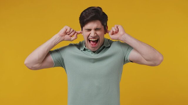 Displeased irritated sad angry fun mad man 20s years old wear green polo shirt closed eyes cover ears do not want to listen scream isolated on plaine light yellow color wall background studio portrait