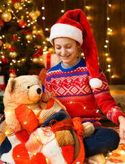 Child girl posing in new year or christmas decoration. Festive lights and lots of gifts, an elegant Christmas tree with toys. The girl is wearing a red sweater and a Santa hat, she playing with bear