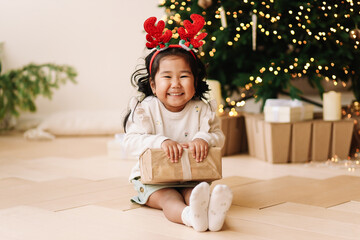 Portrait of a cheerful happy positive emotional Asian little girl the child is having fun rejoicing and holding a gift box on a Christmas holiday in a decorated house during the New Year vacation. 