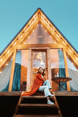 Woman is sitting on the porch of a wooden hut with lights of garlands in the evening. The concept of glamping and renting a chalet. - obrazy, fototapety, plakaty