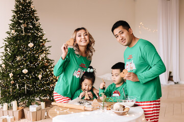 A happy mixed Asian family with two children in green pajamas cook cookies together and prepare for the Christmas holiday on New Year's weekend in a decorated room at home. Selective focus
