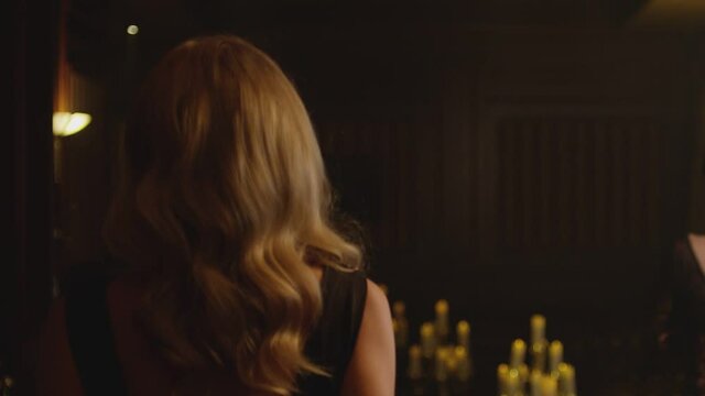 Back view of beautiful blond woman in black elegant dress  walking slowly in dark room with lighted candles . Model walking in luxury fashion show . Expensive home interior . Slow motion . 
