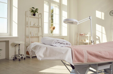 Beautiful interior of empty spa salon, massage room, beauty centre, or dermatologist's office with massage table, modern lamp magnifier, white walls, and shelves with products for skin care procedures
