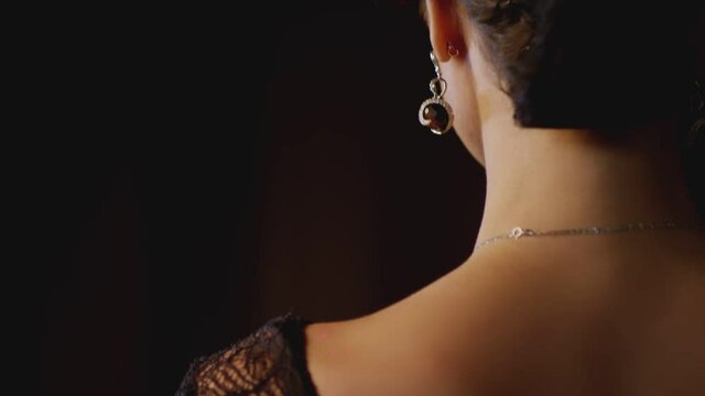 Back view of Beautiful elegant woman in lace or guipure black dress standing , wearing luxury jewelry . Model showing golden necklace and earrings with diamond stones . Black background . Slow motion