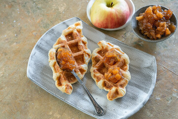 Two waffles of croffle with apple jam are on a ceramic plate.Fresh half of an apple and a saucepan with fruit jam. A popular Korean delicacy..