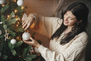 Winter holidays preparations. Happy woman in cozy sweater decorating christmas tree with modern...