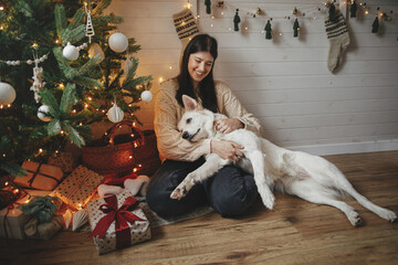 Stylish happy woman hugging adorable dog under christmas tree with gifts and lights. Young female sitting and caressing cute dog in festive scandinavian room. Pet and winter holidays