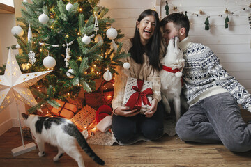 Stylish happy couple having fun with cute dog on background of christmas presents, xmas tree in lights in festive decorated room. Adorable dog kissing family owners in cozy sweaters. Merry Christmas!