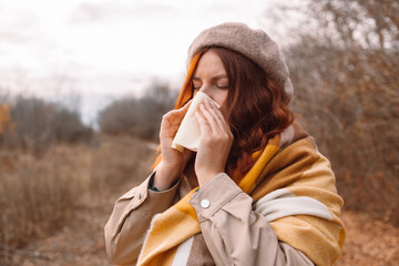 Sick young woman with cold and flu standing outdoors, sneezing, wiping nose with paper napkin in the city autumn park