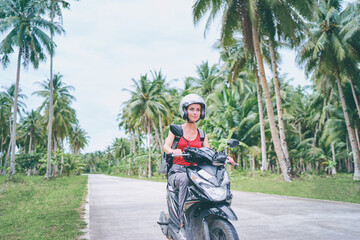 Obraz premium Tropical travel and transport. Happy young woman in helmet riding scooter on the road with palm trees.