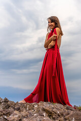 A woman in a red dress stands above a stormy sky, her dress fluttering, the fabric flying in the wind.