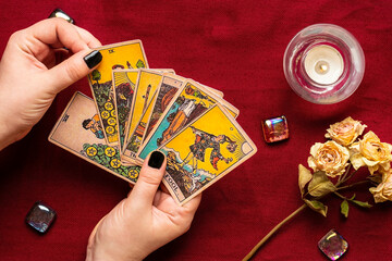 Fortuneteller's hand with black manicure lays out tarot cards, crystal, candle, dry roses on red tablecloth Flat lay Top view Fortune telling, prediction, esoteric concept - 469930583