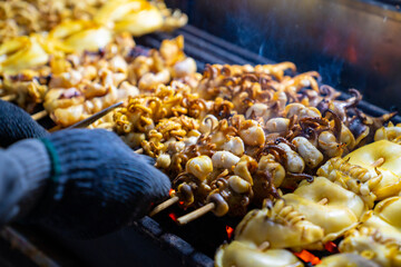 Close up of Chef hand cooking grilled squid on wooden stick with spicy sauce at street night market in Bangkok city, Thailand. Traditional delicious street food grilled seafood on charcoal grill.
