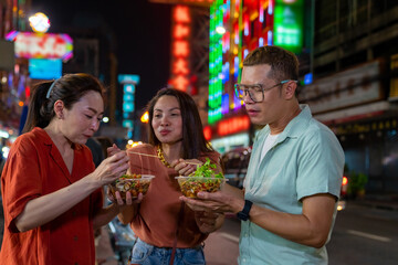 Group of Asian woman and LGBTQ people friends tourist enjoy eating traditional street food bbq seafood grilled squid with spicy sauce together at china town street night market in Bangkok, Thailand