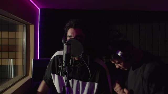 Portrait of inspired stylish handsome multiethnic male hip hop artists in headphones and rapper attire singing into microphone, recording new hit rap song in sound recording studio.
