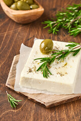 Cheese feta with rosemary, herbs, olives and olive oil on wooden cutting board on old wooden background. Traditional Greek homemade cheese. Selective focus.