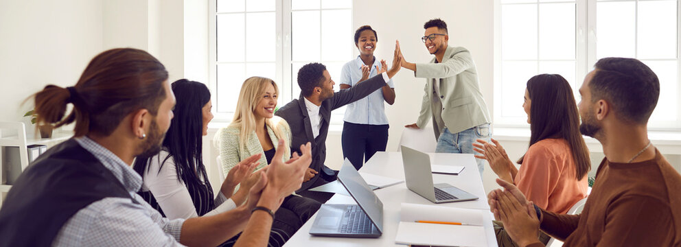 Mixed race business team celebrating good results in office meeting. Happy young black man gives high five to coworker while diverse multi ethnic teammates are applauding. Teamwork and success concept