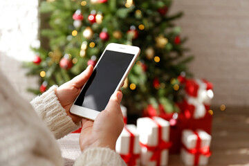 Cropped shot of female hands holding a phone with blank screen over decorated christmas tree and...