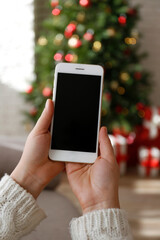 Cropped shot of female hands holding a phone with blank screen over decorated christmas tree and stack of presents under it. First person view. Close up, copy space, background.