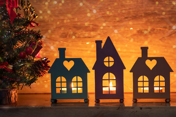 Greeting Christmas card with candles in the candlesticks of the houses and a decorated Christmas tree on the mantelpiece. Festive background with lights in the windows of small houses, bokeh and spark