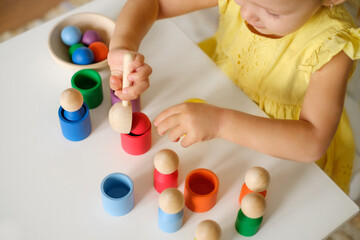 Toddler plays with different figures at the table, sorting them by color, throwing a ball with a spoon into a cup. Early development of a baby using games and toys. Logical thinking and Entertainments - obrazy, fototapety, plakaty