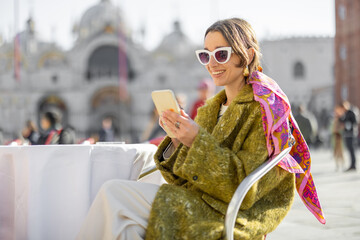 Young woman with phone sitting at outdoor cafe on the famous saint Marks square in Venice. Concept of vacations in Italy. Idea of Italian style and lifestyle. Woman wearing coat and colorful shawl