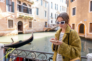 Fototapeta na wymiar Young woman using phone while walking near beautiful water canals in Venice. Concept of vacations in Italy. Caucasian stylish woman wearing coat sunglasses and colorful shawl