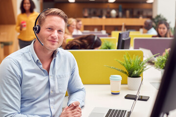 Portrait Of Businessman Wearing Headset Talking To Caller In Busy Customer Services Centre - obrazy, fototapety, plakaty