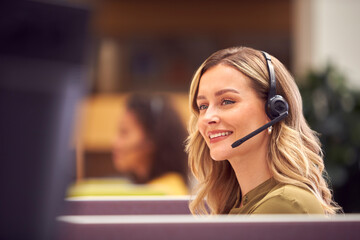 Businesswoman In Cubicle Wearing Headset Talking To Caller In Busy Customer Services Centre