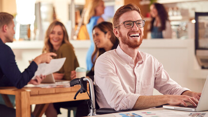 Laughing Businessman In Wheelchair Sitting At Desk Working On Laptop In Busy Office