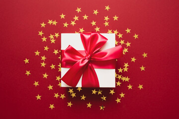 White gift box with ribbon on a red with golden confetti in the form of stars. Valentines, Christmas or New Year day backdrop. Flat lay style with minimalistic design.