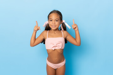 Happy little girl in swimwear pointing fingers up standing isolated over blue background.