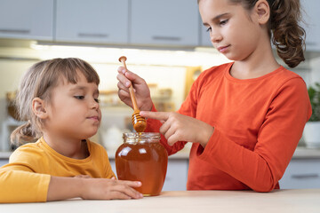 Happy children eating honey from a jar at home in the kitchen. funny girls lick honey from a spoon. Healthy organic food sweets for kids