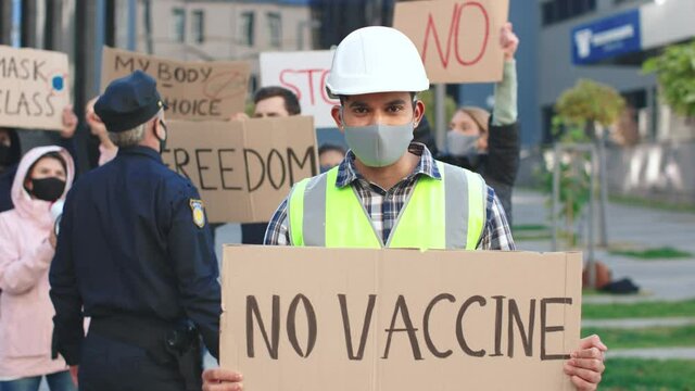 Caucasian builder or architect wearing protective mask and helmet standing at the street and holding placard no vaccine. People with placards and posters on public demonstration, no covid 19 vaccine