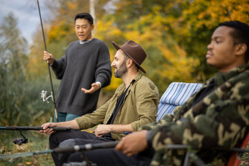 Asian man talking to male multiracial friends during fishing in nature. Men resting and spending time together on river or lake shore. Concept of leisure, hobby and weekend in nature. Friendship