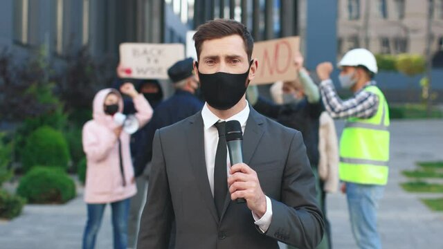 Male Journalist Correspondent Wearing Suit Reporting Live Near Building. People With Placards And Posters On Public Demonstration, No Covid 19 Vaccine Concept