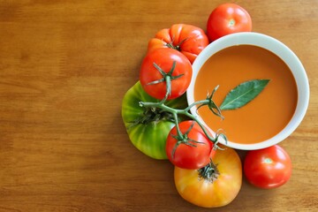 Bowl of tomato soup garnished with bay leaf surrounded by various tomatoes on wood background with copy space.