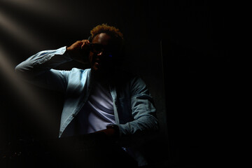 African-American DJ with headphones is sitting in a dark room, small rays of light fall on him. DJ...