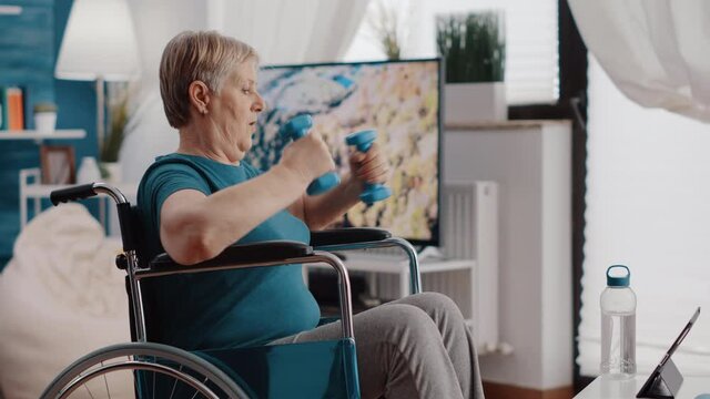 Old woman with disability using dumbbells to exercise and watching workout video on digital tablet. Retired person sitting in wheelchair, training with weights and looking at online lesson