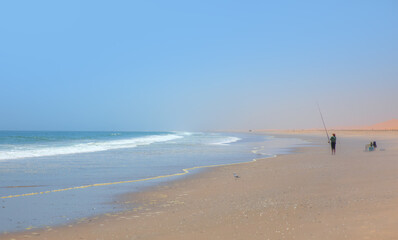 Fisherman fighting fish with a long fishing rod on Atlantic ocean beach in front of beautiful blue sky