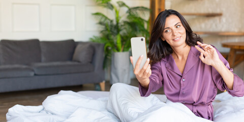 Middle aged woman lies in bed with cell phone. She wake up make selfie for social media posts. Modern technology and digital addiction concept. Format photo 2x1.